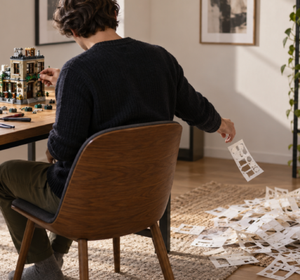 A hobbyist builds with bricks at a table while a large pile of empty sticker-sheet backings lies on the floor and one backing drops from their hand.