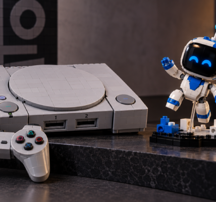 A brick-built retro game console inspired by the original PlayStation sits beside a small white-and-blue robot collectible on a dark tabletop.