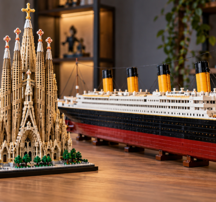 A detailed brick-built cathedral model stands beside a much longer ocean-liner-style model on a wooden tabletop.