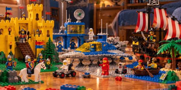 A nostalgic playroom floor scene combining a yellow medieval castle with knights, classic space builds with astronauts, and a pirate ship and island.